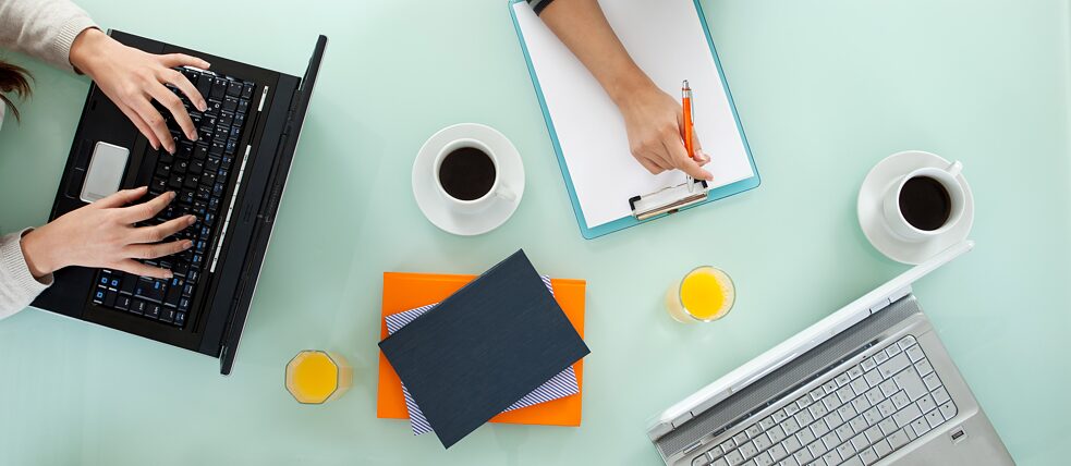 Bir masanın üstten görünümü.  Masada iki laptop, iki kahve fincanı, iki bardak, portakal suyu ve defterler var. Bir kişi not alıyor, biri laptopta yazıyor.