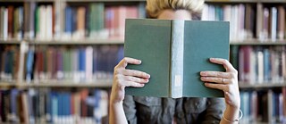 Eine Frau liest in einem Buch, ihr Gesicht ist vom Buch verdeckt. Im Hintergrund stehen Bücherregale.