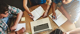 Blick von oben auf junge Leute in einem Café. Auf dem Tisch sind Laptop, Smartphone und Notizbücher.