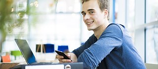Ein junger Mann sitzt in einem Café. Vor ihm steht ein Laptop. Er hält ein Handy in der Hand und lächelt.