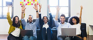 Junge Leute sitzen mit Laptops vor einer Couch auf dem Boden. Sie werfen jubelnd die Arme in die Höhe.