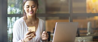 Eine junge Frau sitzt im Büro an einem Tisch und blickt auf ein Handy. Vor ihr steht ein Laptop.