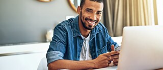 Ein junger Mann sitzt an einem Tisch. Vor ihm steht ein Laptop.