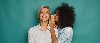 Zwei junge Frauen stehen vor einer türkisfarbenen Wand. Die eine flüstert hinter vorgehaltener Hand der anderen etwas ins Ohr. Diese lacht.