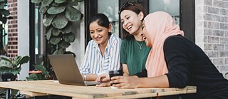 ثلاث سيدات يجلسن حول طاولة في الهواء الطلق، وأمامهن جهاز لاب توب. يبدو أنهن يعقدن محادثة عبر الهاتف. 