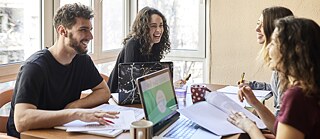 Vier junge Leute sitzen um einen Tisch. Vor ihnen stehen Laptops. Sie arbeiten vergnügt an Notizen.