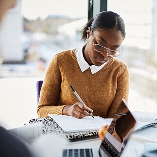 Eine junge Frau lernt konzentriert, vor ihr liegen viele Arbeitsmaterialien. Im Hintergrund ist eine moderne Stadtansicht unscharf zu sehen.