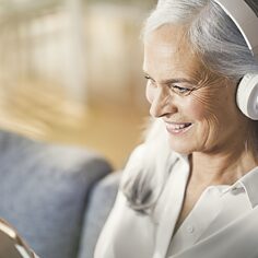 Eine Best-Agerin sitzt auf einer Couch. Sie hat Kopfhörer auf und blickt auf ein Tablet.