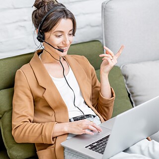 A young woman is sitting on a sofa. She is taking part in an online meeting and wearing a headset.