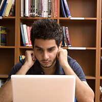 Ein junger Mann liest konzentriert in seinem Laptop. Im Hintergrund steht ein Bücherregal.