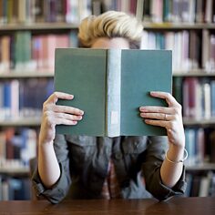 Eine Frau liest in einem Buch, ihr Gesicht ist vom Buch verdeckt. Im Hintergrund stehen Bücherregale.