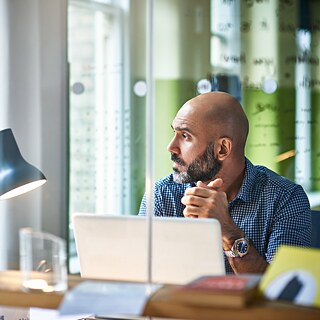 Ein Geschäftsmann sitzt im Büro am Schreibtisch und blickt nachdenklich aus dem Fenster.