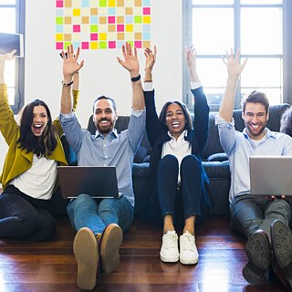 Junge Leute sitzen mit Laptops vor einer Couch auf dem Boden. Sie werfen jubelnd die Arme in die Höhe.