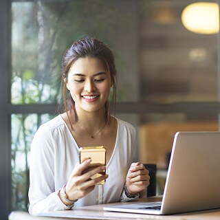 Eine junge Frau sitzt im Büro an einem Tisch und blickt auf ein Handy. Vor ihr steht ein Laptop.