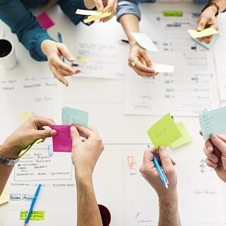 Blick von oben auf einen Tisch mit Papieren und Notizen. Mehrere Hände halten bunte Post-Its.