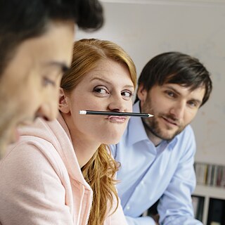 Drei junge Menschen scherzen. Eine junge Frau hält einen Bleistift mit der Oberlippe unter der Nase als „Schnurrbart“.