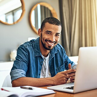 Ein junger Mann sitzt an einem Tisch. Vor ihm steht ein Laptop.