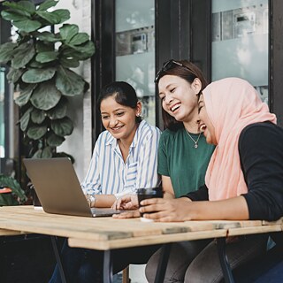 Drei Frauen sitzen im Freien an einem Tisch. Vor ihnen steht ein Laptop. Sie halten vermutlich eine Telefonkonferenz ab.