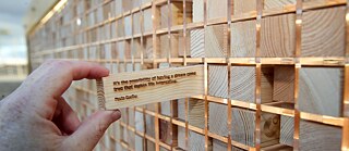 A hand pulls a small wooden block out of a wall that is built out of nothing but small wooden blocks. On the block, the following writing is engraved: "It's the possibility of having a dream come true that makes life interesting. - Paulo Coelho"