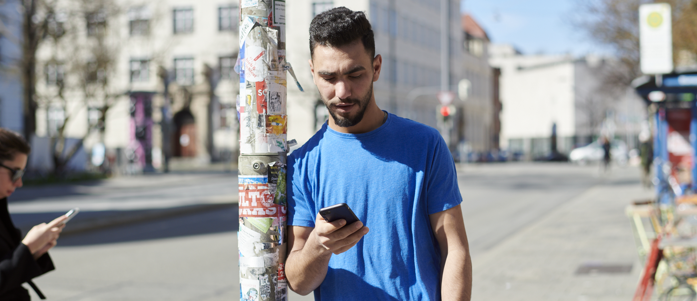 Ein junger Mann lehnt an einer Straßenlaterne und schaut auf sein Smartphone.