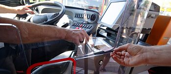 Eine Hand reicht einen Geldschein durch eine Plexiglasscheibe, die den Fahrstand eines Busses vom Fahrgastraum trennt. 