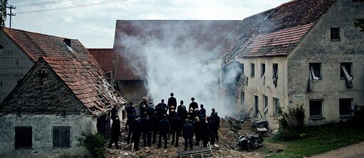 Schwarz gekleidete Menschen stehen vor der Ruine eines rauchenden Bauernhofs.