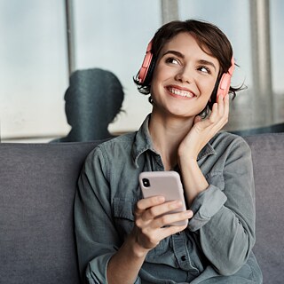 Ein junges lächelndes Mädchen mit rosa Kopfhörern sitzt auf einer Couch und hört etwas auf ihrem Handy.