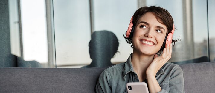 A young smiling girl with pink headphones sits on a couch listening to something on her mobile phone.
