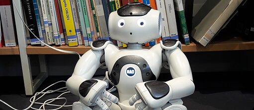 Ein Roboter vom Typ NAO 6 sitzt cvor einem Bücherregal
