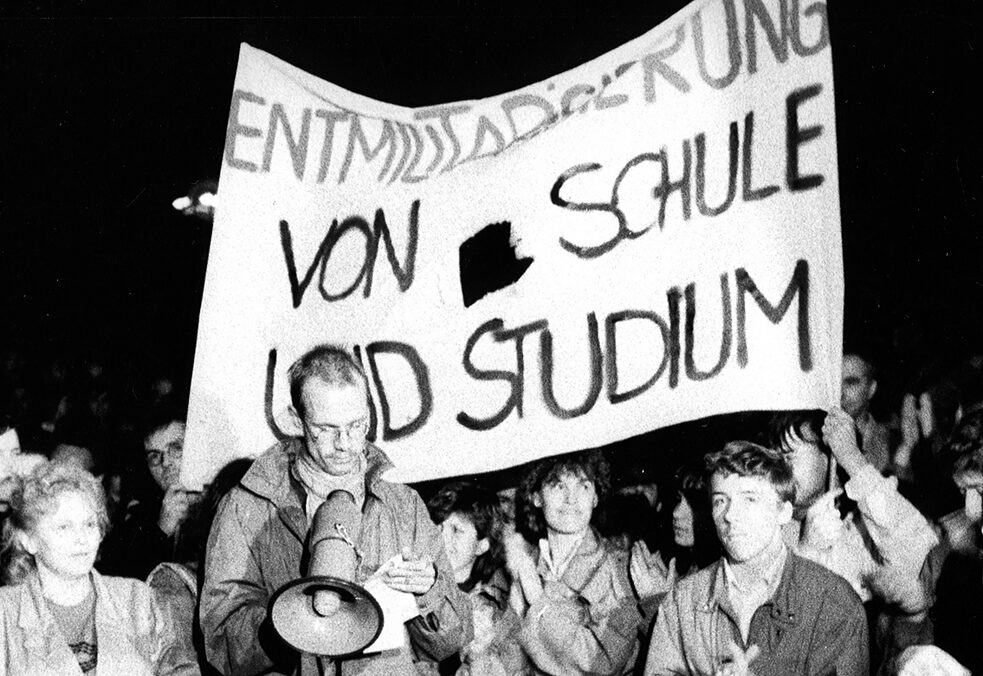 Demonstratsioonist osavõtjad seisavad, plaksutavad ja vaatavad kaamera poole. Kaks neist hoiavad käes suurt loosungit kirjaga „Entmilitarisierung von Schule und Studium“ („Kooli ja ülikooliõpingute demilitariseerimine“)