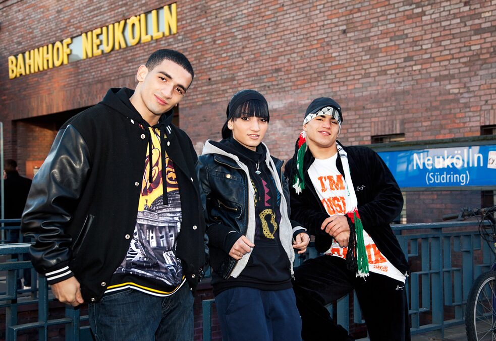 Die drei Geschwister Hassan, Lial und Maradona lehnen an einem Geländer vor dem Bahnhof Neukölln. Sie blicken freundlich in die Kamera.