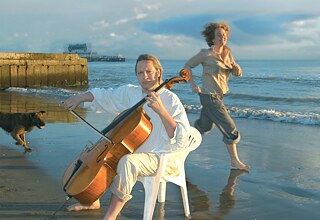 Ein Mann spielt am Strand Cello. Hinter ihm läuft eine Frau, die lächelt. 