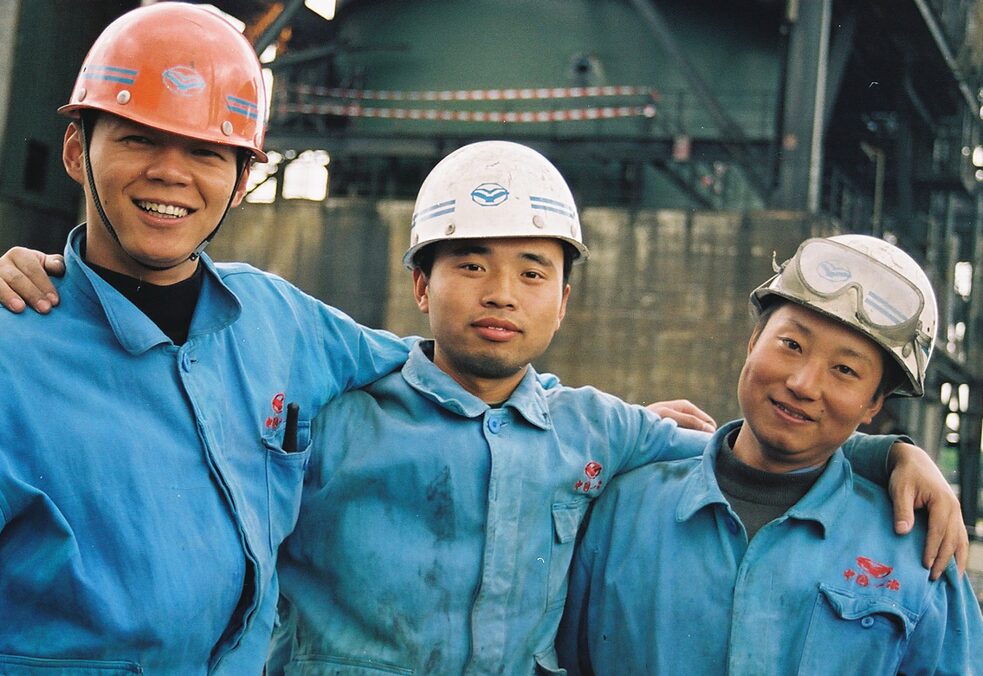 Drei junge Chinesen in Arbeiteranzügen und mit Helmen stehen nebeneinander und lächeln in die Kamera. 