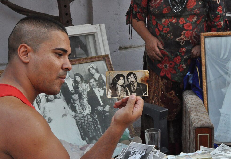 Ein junger Mann hält eine alte Fotografie eines Paares in die Kamera. Im Hintergrund sind Hochzeitsfotos zu sehen.