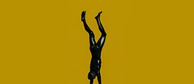Siyah bronz bir erkek heykeli, sarı bir fon önünde amuda kalkıyor.