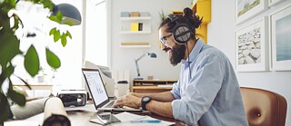Ein junger Mann sitzt in einem hellen, freundlichen Büro am Schreibtisch und tippt in ein Laptop.