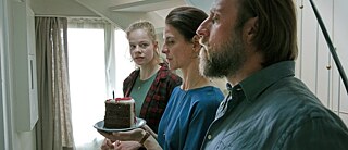 Trois personnes devant une porte fermée avec un gâteau d'anniversaire