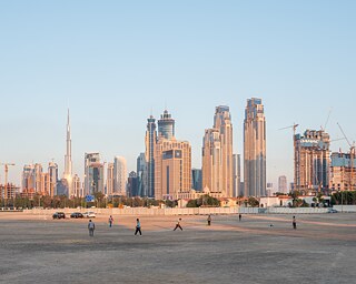 رجال يلعبون الكريكيت في أحد الأماكن القليلة المفتوحة في منطقة جُميرة، وفي الخلفية يظهر خط أفق مدينة دبي. المرافق الرياضية غير متوفرة للطبقة العاملة، على الرغم من الرفاهية في دبي، كما أن هناك أماكن عامة قليلة للمارسة الرياضة، مما يؤدي إلى ارتجال ملاعب للمارسة مبارة للكريكيت عندما تسنح الفرصة.