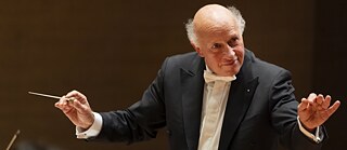 Der Dirigent Wolfgang Christ hält einen Taktstock und blickt nach links auf sein Orchester, welches nicht im Bild ist.