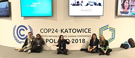 5 young activists pose in front of the logo of the COP24. There are several screens to be seen above them.