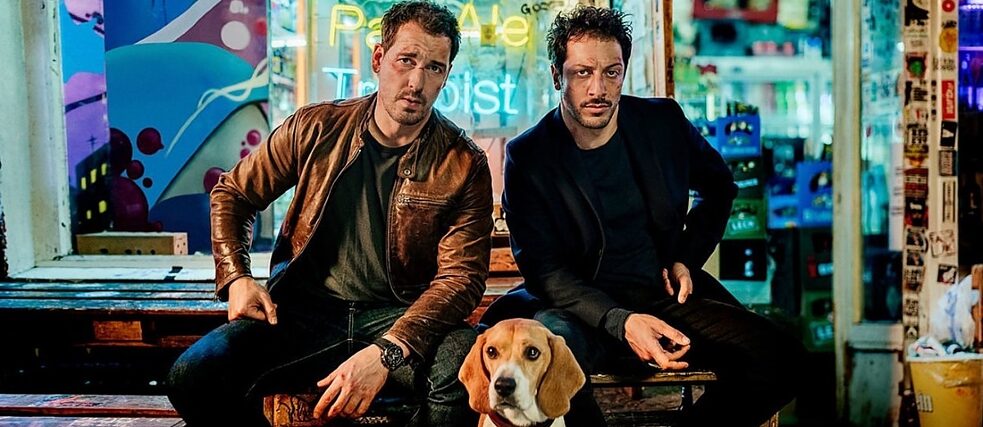 Still frame from the Netflix Series „Dogs of Berlin“: Felix Kramer as Kurt Grimmer and Fahri Yardim as Erol Birkan sitting in front of a colorful Berliner convenience store.
