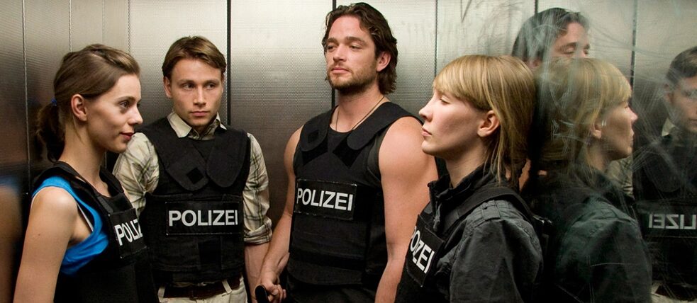 Standbild aus der Serie „Im Angesicht des Verbrechens“: Carmen Birk, Klara Manzel, Max Riemelt, Ronald Zehrfeld in Polizei Schutzwesten stehend in einem Aufzug