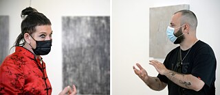 A collage of two photos. On the right photo, a man with shaved head and tattoos on both arms stands in front of a white wall on which a metallic artwork is hanging. He wears a facemask and stretches his hands in front of his breast. On the left photo a middle-aged woman in red blouse with a topknot and a face mask stands in front of two grey artworks and stares at something.