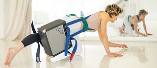 Eine Frau mit angebundenem Gitarrenverstärker bewegt sich auf Füßen und Händen nah am Boden