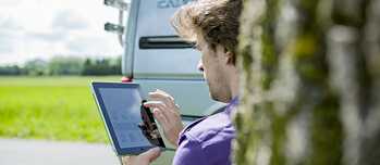 Rückansicht auf einen Mann, der neben einem Camping-Bus an einem Baum lehnt. Er hält ein Tablet in der Hand und sieht konzentriert darauf. Im Hintergrund sieht man die Straße. 