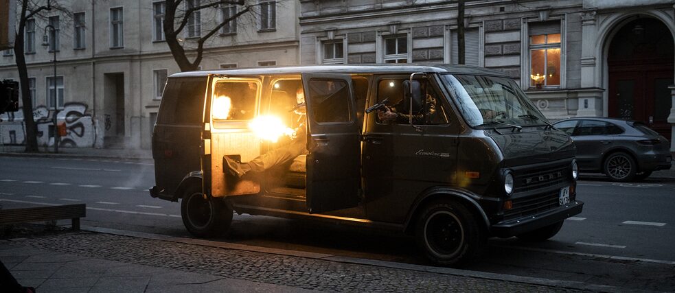 Still from the Netflix series "Dogs of Berlin": There is drive-by shooting from a panel van out in the open street, muzzle flashes and weapons from the van are aimed at an unseen target.