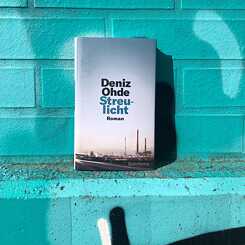 Das Buchcover zu Deniz Ohdes Roman "Streulicht" vor einer türkisfarbenen Hauswand.