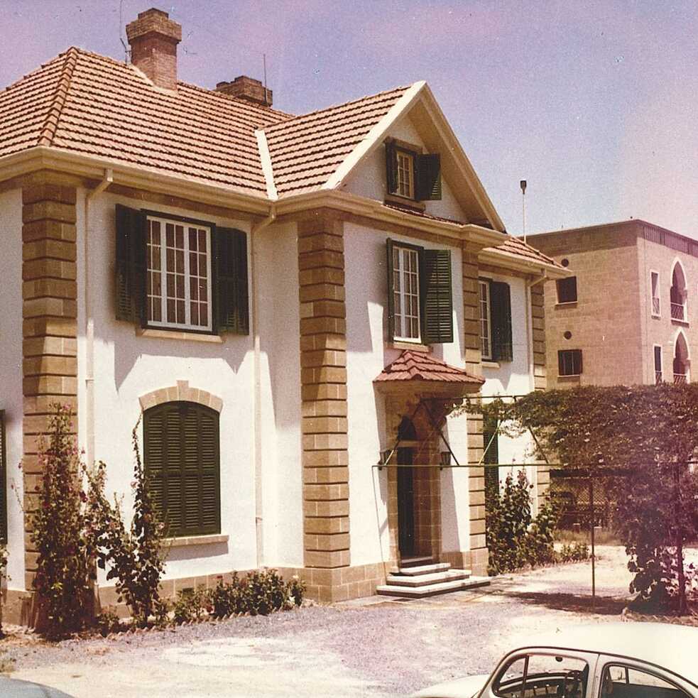 Το κτίριο του Ινστιτούτου Γκαίτε κάτω από τον γαλάζιο ουρανό. Ο τοίχος του κτιρίου είναι άσπρος με πουρόπετρα στις γωνιές. Τα παραθυρόφυλλα είναι σκούρο πράσινο. Υπάρχουν τρία αυτοκίνητα στον χώρο στάθμευσής μπροστά από το κτίριο. Στο βάθος φαίνεται το Ξενοδοχείο Λήδρα Πάλας.