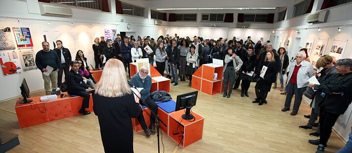 At the microphone is Karin Varga, Director of the Goethe-Institut Cyprus, who gives an opening speech and stands with her back to the camera. The visitors sit on orange benches, which belong to an installation of the exhibition, or stand spread out in the room. They listen to the speech and sometimes hold brochures of the exhibition in their hands. Posters on the theme of Bauhaus hang on the walls, as does the exhibition on the work of the Cypriot architect Fivos Polydorides. The latter is standing on the right of the picture and is wearing a red pullover and a white shirt. His hair is white and he wears glasses.