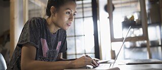 Eine Frau sitzt im Büro am Schreibtisch und arbeitet konzentriert am Laptop.